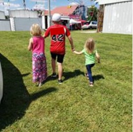 3 kids, the tallest in the center, the shortest on the right, walk away from the camera. They are outside on a sunny day.